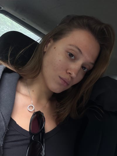 A young woman with brown hair and fair skin is photographed from the chest up, looking directly at the camera. She is wearing a dark ribbed top and a grey jacket. A silver necklace with a circular pendant adorns her neck. Sunglasses are clipped to the front of her shirt. The background suggests she is inside a vehicle, with a blurred window and dark upholstery visible. The lighting is natural, coming from an unseen source to her right. The overall impression is a candid, informal portrait.