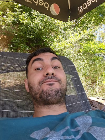 A medium shot of a fair-skinned man lying on a chaise lounge under a Pepsi umbrella. The man has dark hair and a beard. He is wearing a teal shirt with a grey graphic. The chaise lounge is dark grey and striped. Lush greenery surrounds the lounge. The location is presumed to be in Slavovitsa, Bulgaria.