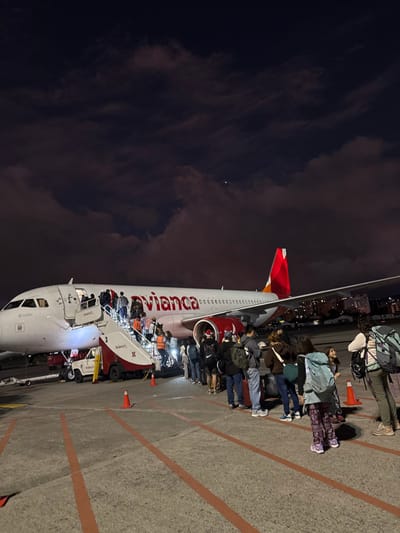 An Avianca airplane stands on the tarmac under a cloudy night sky. Passengers are boarding via a mobile staircase attached to the front of the plane. People are queuing on the ground, waiting to board, and others can be seen ascending the stairs. The plane features the Avianca logo prominently, with a red tail fin. The surrounding environment includes airport markings and some distant city lights.