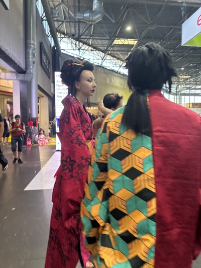 Two individuals are in the foreground of a well-lit indoor event space, likely a convention center. The person on the left, viewed from the side, is wearing a vibrant red kimono with black floral patterns and has an elaborate updo hairstyle with red accents. This person has dramatic eye makeup. The person on the right is viewed from behind and wears a garment that is half plain red and half patterned with a geometric design in teal, yellow, and black. They have dark hair styled in a ponytail that extends down their back. In the background, other attendees are visible, some browsing displays or walking. Signage for "Halls 5-6" is present on a wall in the mid-ground, indicating this is likely the Paris-Nord Villepinte Exhibition Centre in Villepinte, France, a common venue for large-scale events and conventions. The floor is a light gray, and the ceiling features exposed industrial beams and ventilation systems.