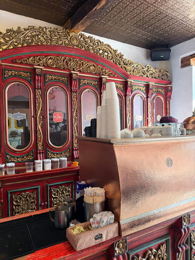 An ornate, partially visible dark-red and gold cabinet features multiple glass-windowed sections and decorative trim. A stack of white disposable cups sits atop the cabinet, with a metallic copper dispenser alongside. Additional items, including sugar packets, stirrers, and containers, are affixed to the cabinet's side. The cabinet is positioned against a wall with textured, brown-toned wallpaper, and a black speaker is visible in the upper-left corner.