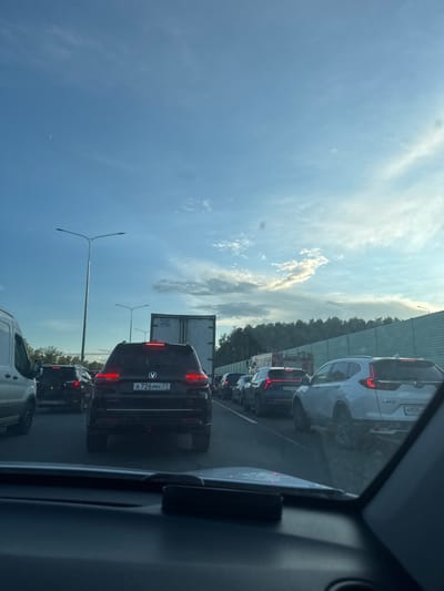 The image depicts a traffic jam on a highway, likely near Krutitsy, Russia, given the presence of Russian license plates. The perspective is from inside a vehicle, looking forward. Several cars are visible in multiple lanes, with brake lights illuminated, indicating slow or stopped movement. The sky is a clear blue with scattered clouds and a crescent moon. Trees and a sound barrier line the side of the highway. Streetlights are also present. The overall scene suggests a typical urban or suburban commute during daylight hours.