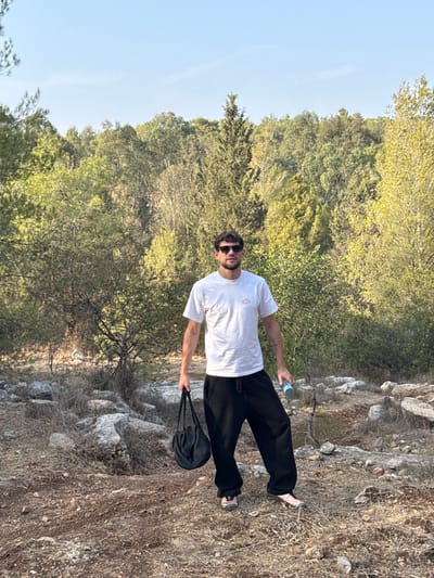 A male individual is standing on an uneven, dry, and rocky path within a natural, forested area of Modi'in-Maccabim-Re'ut, Israel. The subject, estimated to be in his late 20s or early 30s, is centered in the frame, facing directly forward. He is wearing dark sunglasses, a light-colored (possibly off-white) short-sleeved t-shirt with a small, light pink graphic on the left chest, loose-fitting black sweatpants, and light-colored sneakers. In his left hand, he holds a black fabric drawstring bag, while his right hand holds a light blue cylindrical object, possibly a water bottle or spray can. The immediate foreground consists of dry, reddish-brown earth, sparse dry vegetation, and scattered light grey stones and rocks. The background features a dense, verdant forest with a variety of tree species, displaying shades of green and some lighter yellowish-green foliage under a clear, light blue sky.