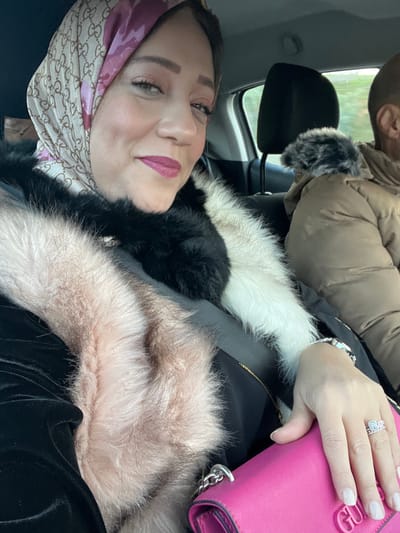 The image features a woman seated inside a car, likely as a passenger, captured in a close-up, angled shot. She is looking towards the camera with a gentle smile, wearing a light pink or beige headscarf patterned with the interlocking "GG" logo, characteristic of Gucci. Her makeup includes winged eyeliner, defined brows, and pink lipstick.

She is dressed in a multi-toned fur coat, exhibiting sections of black, white, and light brown or pinkish fur. Her right hand, holding a bright pink handbag with the embossed "GUESS" logo, is visible. On her right ring finger, she wears a silver ring with a decorative, possibly geometric, design. A silver watch is partly visible on her left wrist.

In the upper background, another person is partially seen from behind, wearing a brown puffer jacket with a fur-trimmed hood, suggesting cooler weather. The setting is the interior of a car, with black upholstery and headrests visible. A blurred outdoor scene, possibly featuring greenery, can be glimpsed through the window, indicating daylight. The overall atmosphere appears relaxed. The location context for the image is Majmaa Tolba, Morocco.