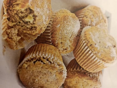 The image displays a close-up, overhead view of several freshly baked muffins. The muffins are golden brown with a slightly crumbly texture and are dusted with powdered sugar. They are arranged in a basket or container, with some muffins overlapping others. The lighting is soft, highlighting the baked goods. There is no discernible indication of the city of Rumaithiya, Kuwait, or any other specific location within the image.