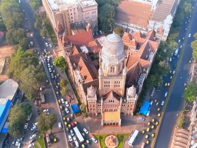 This aerial image captures a busy street scene in front of a grand, ornate building with multiple domes and towers, likely a historical or governmental structure. The building's architecture features intricate stonework, arched windows, and a large central dome. The street is filled with traffic, primarily consisting of yellow auto-rickshaws and white cars, indicating a bustling urban environment. A white bus is also visible, suggesting public transportation is in use. The presence of trees lining the street and surrounding the building indicates a well-established area, possibly a park or a historical district. The lighting suggests it is daytime, with the sun casting shadows, but the exact time of day is not definitively clear. There is no visible text within the image. The scene conveys a sense of energetic activity and movement due to the volume of vehicles and the implied presence of people going about their daily routines. The image provides a high-level view, emphasizing the scale of the building and the density of the surrounding urban infrastructure.