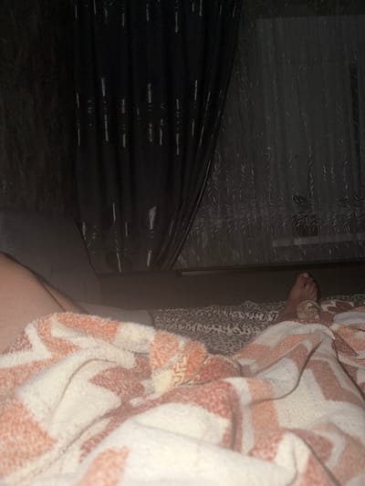 The image depicts a dimly lit bedroom scene. In the foreground, a person's legs are covered by a soft, patterned blanket in shades of white and peach. The blanket has a zigzag design. To the right, a bare foot is visible resting on the bed. The bed is covered with patterned sheets, possibly a leopard print. In the background, dark curtains hang over a window, with sheer, patterned curtains visible underneath. The overall impression is that of a private, domestic space. The location is identified as a bedroom in the city of Avdarma, Moldova.
