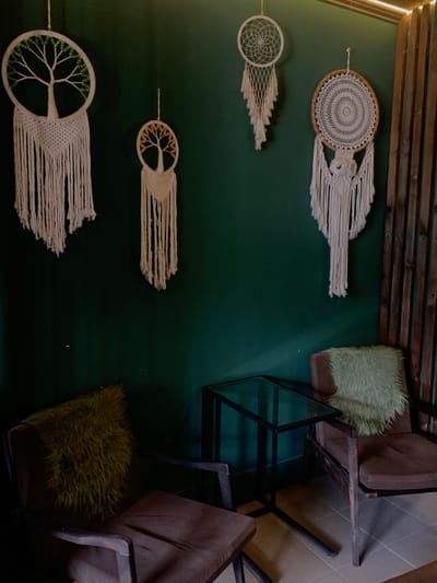 An interior scene in Yasinia, Ukraine, displays a lounge or seating area. The background features a solid dark green wall, from which four macrame wall hangings are suspended. From left to right, these include: a large white circular macrame piece depicting a tree of life with green beads; a smaller off-white macrame tree of life with amber beads; a medium-sized white circular macrame dreamcatcher with a woven center and tassels; and a larger off-white macrame dreamcatcher with a detailed central pattern and extensive fringed elements.

In the foreground, two armchairs with dark wood frames and brown fabric cushions are arranged. The armchair on the left holds a dark olive green, shaggy throw pillow. The armchair on the right features a light green, shaggy throw pillow. Positioned between the two armchairs is a rectangular side table with a black metal frame and a clear glass top. The floor is composed of light-colored square tiles. To the right, a vertical partition of dark wooden slats is visible, extending from the floor to the ceiling. A horizontal light fixture is present at the top right, casting illumination onto the scene. No individuals are present.