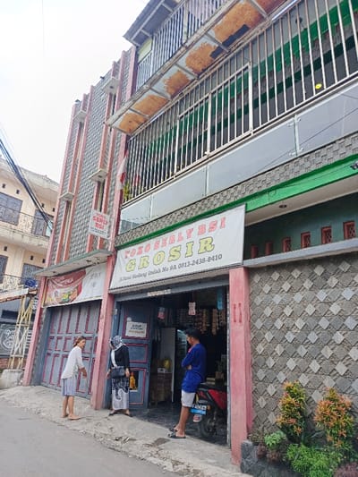 A building with two visible stories, featuring a shop named "Toko Serly BSI Grosir" on the ground floor. The shop front is open, with three people present. A woman in a white top and shorts stands to the left, another woman in a headscarf stands next to her, and a man in a blue shirt leans against the building on the right, near a parked motorcycle. The building is constructed with brickwork and stone patterns, with balconies on the upper levels.