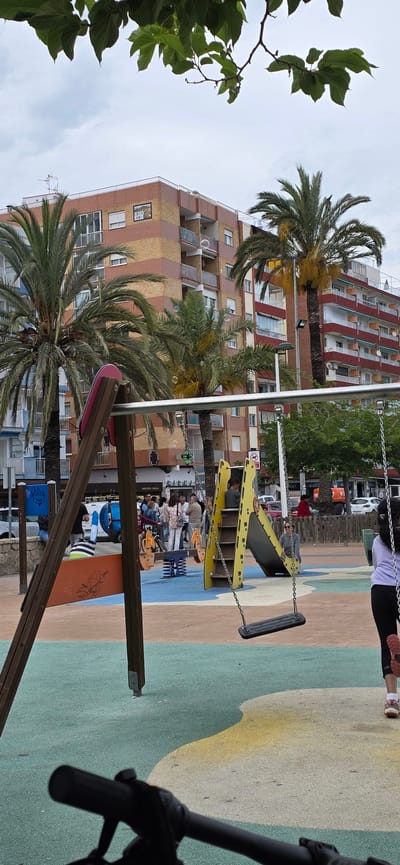 The image shows a playground in El Port De Sagunt, Spain. In the background is a tall building with a brown and orange striped facade, interspersed with palm trees. There is a yellow and gray slide with a metal ladder, a swing set with a black seat, and other playground equipment. People, including children and adults, are scattered around the playground. Some are on the equipment, while others are standing or sitting nearby. The playground surface is painted in various colors, including green, beige, and blue. A tree with green leaves is on the left side of the image. Part of a bicycle handlebar is visible in the foreground on the right.