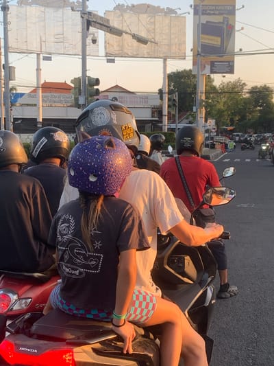 This is a medium shot, eye-level, slightly angled, outdoor image taken in Denpasar Barat, Indonesia, during the golden hour. The scene depicts a traffic intersection with multiple people riding on scooters. In the foreground, a young female with dark hair braided down her back sits on the back of a scooter, wearing a dark t-shirt with a graphic design and checkered shorts. She is wearing a distinctive purple helmet adorned with leopard print and cat-like ears. Next to her on the same scooter, a person wearing a light-colored t-shirt and dark shorts is visible. Another scooter in the immediate foreground has a red body and the "HONDA" logo is visible on its rear.
Behind the foreground scooters, several other individuals are riding. One person wears a red t-shirt and a black helmet with white lettering that reads "PURE IRON TATTOO STUDIO." Another person to their left wears a dark helmet with a sticker reading "MERDAHBALI." Further back, more scooters and their riders are partially visible, some wearing helmets.
The environment includes traffic lights, utility poles, overhead signs, and billboards. One billboard displays an advertisement for "MODERN KRETEK" and "DUNHILL." Another sign appears to be for a hotel or resort, reading "Feel it cozy like at Home Pet Friendly Best." The road is asphalt, and a pedestrian crossing is marked with white stripes. Trees and buildings line the background. The overall impression is a busy street scene in Denpasar Barat during sunset.