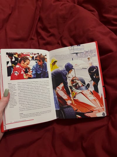 This is a photograph of an open book lying on a red textured surface, likely fabric. The book's pages feature text and images related to motorsports.

On the left page, a black and white photograph shows two men. The man on the left is wearing a red racing suit with the number 3 prominently displayed on it. He is facing the man on the right, who is wearing a blue denim shirt and sunglasses.

The right page contains a color photograph depicting a red Formula 1 race car on a track. A man in a racing suit and helmet is seated in the car. Several people are gathered around the car, some appearing to be mechanics or team personnel. One person is holding an umbrella over the driver. The environment appears to be a race track with spectators in the background. A large "SEIKO" sign is visible above the track.

The book also contains Russian text, with headings such as "Вверх" (Up), "Справка" (Reference), and "Возвращение на вершину" (Return to the top). The number "74" is visible on the bottom left of the page.

The image does not contain any information about the city of Vysokaya Gora, Russia.