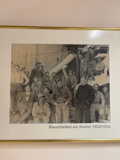 This is a black and white photograph of a group of approximately 20 men posing outdoors, likely at a construction site given the background which features rough wooden structures and debris. The men are dressed in what appears to be work attire, with many wearing hats and suspenders. Some men are seated, while others stand, with the majority looking towards the camera. In the foreground, one man is dressed in a suit, appearing to be in a supervisory role. The photograph is framed and mounted on a wall, with a printed caption below it that reads "Bauarbeiten am Kamm 1924/1926". The text below the image indicates that the location is Ehrwald, Austria.