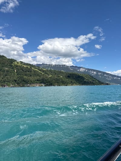 This image captures a scenic view of the city of Oberhofen am Thunersee, Switzerland, from the perspective of a body of water, likely Lake Thun. The foreground is dominated by the turquoise water of the lake, with gentle waves and white foam indicating movement.
In the midground, a picturesque village lines the shore. Numerous buildings with red-tiled roofs are nestled amongst lush green trees that climb up the steep hillside. Several boats are visible docked along the shoreline, suggesting a popular tourist destination.
The background features the dramatic mountainous landscape characteristic of the Swiss Alps. The mountains are covered in dense green forests on their lower slopes, transitioning to rugged, rocky faces higher up. The sky above is a vibrant blue, dotted with fluffy white cumulus clouds, indicating fair weather. The overall impression is one of natural beauty and serene lakeside living.