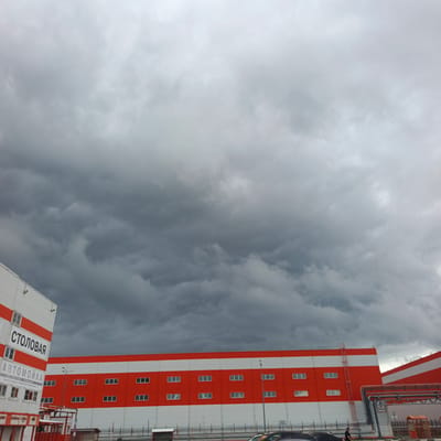 This image depicts an overcast day in Sof'ino, Russia, with a dramatic sky filled with dark, heavy clouds that suggest impending rain. In the foreground, two industrial-style buildings are visible, one prominently featuring white and bright orange horizontal banding. The building on the left has large text on its facade that reads "СТОЛОВАЯ" (Cafeteria) and "АВТОМОЙКА" (Car Wash). Below "АВТОМОЙКА," there is smaller text including a phone number. Several windows are visible on the buildings.

A person is seen walking near the base of the building on the right, near some industrial piping. The scene appears to be a commercial or industrial area, possibly a business park. The lighting is dim due to the heavy cloud cover, indicating it's likely daytime but not sunny. The overall mood is somber, dictated by the weather and the imposing architecture. The visible text provides clues about the services offered at the location.