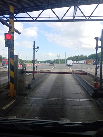 The image depicts a view from inside a vehicle approaching a toll booth on a highway. The toll booth has a red light illuminated, indicating a stop. A red barrier arm is lowered across the lane. In the background, other vehicles, including trucks and cars, are visible on the highway. The environment appears to be an outdoor setting with a cloudy sky. Based on visual cues consistent with infrastructure and vehicle types commonly found in Poland, this scene is likely located at a toll plaza on a highway near Mysłowice, Poland.