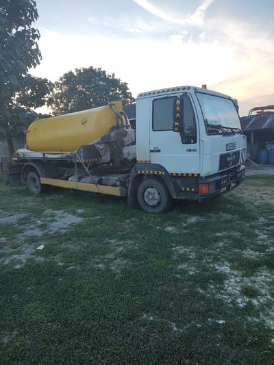 In the city of Kochmar, Bulgaria, a white MAN truck with a yellow tank is parked on a grassy area. The truck has reflective yellow and black chevron markings on its sides and front. A yellow barrel-shaped tank is mounted on the truck's chassis. The truck appears to be used for hauling liquids or waste. In the background, there are trees and a rustic building with a corrugated metal roof. The sky is partly cloudy with a hint of sunset colors. The overall scene suggests a rural or industrial setting.