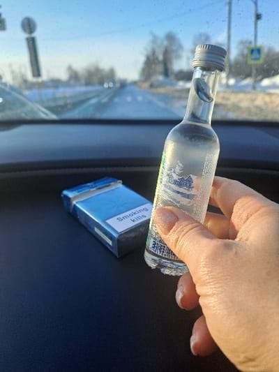 A hand holds a small, nearly empty bottle of clear liquid, likely alcohol. The bottle lies horizontally above a blue cigarette pack labeled "Smoking Kills." These items rest on a dark, possibly leather surface inside a vehicle. Outside the windshield, a road and wintery landscape are visible under a bright sky. The scene suggests the potential for irresponsible or dangerous behavior.