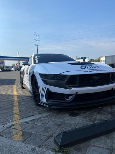 A white Ford Mustang with black decals is parked on a paved surface, likely a parking lot or roadside. The car has prominent "Ford" lettering on the hood and "www.raceworkshop.com" as well. The location appears to be at a gas station, identified by the gas pumps and pricing boards. The photo was taken during daylight hours with a clear, blue sky in the background. Power lines and utility poles are also visible in the background, indicating an urban or semi-urban setting. The presence of other parked cars suggests a public area. No people are visible in the immediate foreground. The exact location is Santa Ana Xalmimilulco, Mexico.
