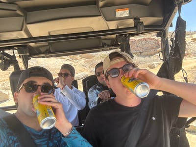 A ground-level shot from inside the open roof of an all-terrain vehicle (ATV) shows four people, seemingly young men, drinking from yellow beverage cans while outside in the city of El Leonero, Mexico. The cans are labeled with a brand name "VOL" and the text "LARA", which is also partially readable, and contain text that states "4,5%". One man has a silver bracelet on his left wrist. One has a white, long-sleeved shirt. The terrain outside features low, brick-type walls and dusty-colored ground. A warning label is visible on the ATV's interior, advising against excessive speed and providing other safety information. The shot is taken at an angle, making the subjects appear upside down.