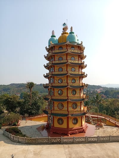 This image depicts a multi-tiered pagoda in Thiện Chí, Vietnam, under a clear, bright blue sky. The pagoda, consisting of at least eight visible levels, is predominantly yellow and red, with green-tiled, curved roofs on each tier, accented by golden trim and ornate detailing. Each level features multiple circular windows or decorative panels, some containing imagery of figures. The top of the structure is crowned with multiple domes, including prominent yellow and turquoise ones, each surmounted by an upward-pointing hand sculpture and a finial.
The pagoda stands on a multi-level concrete plaza, partially bordered by a carved stone railing. A reddish-brown tiled platform directly surrounds the pagoda's base. To the right of the pagoda, a red pedestrian bridge with golden railings curves away from the structure, near a decorative planter containing red flowers. The surrounding environment consists of lush green trees, with rolling hills and scattered residential or small commercial buildings visible in the distant background. The scene is brightly lit by direct sunlight. No people are visible.