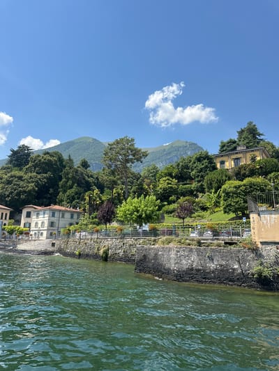 This is a daytime, eye-level, outdoor, horizontal, medium shot of a lake in Tremezzina, Italy. The water in the foreground is a dark green, with ripples and sunlight glinting off its surface. In the midground, a stone embankment lines the shore, with a decorative metal fence and a walkway behind it. Several cars are parked along the walkway. Behind the walkway, a lush green hillside rises, dotted with trees and several buildings. One building, a pale yellow villa with a red-tiled roof, is prominent on the hillside, bearing a sign that reads "VILLA GELSOMINO". Another lighter-colored building with a similar roof is visible to the left, with stairs leading down to the water. The hillside is densely vegetated with various shades of green trees and shrubs. In the background, a large, green mountain range dominates the horizon, with a clear blue sky above, featuring a few scattered white clouds. The overall scene depicts a serene lakeside view of Tremezzina.