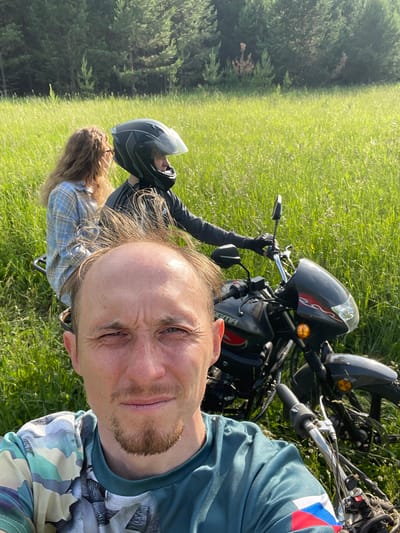 In the village of Nizhnii Bugrysh, Russia, a group of individuals is captured in a selfie-style photograph. In the foreground, a man with short, light brown hair and a goatee stares at the camera with a concerned expression. He wears a camouflage t-shirt. Behind him, a motorcycle is partially visible, with a rider wearing a black helmet and a dark jacket and a passenger wearing a blue plaid shirt. The motorcycle appears to be parked in a field of tall green grass, with a forest in the background. The overall scene suggests an outdoor excursion.