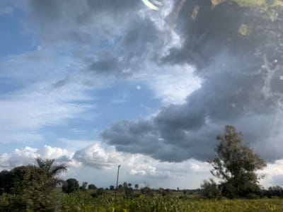 An overcast sky with large, dark storm clouds dominates the upper portion of the image. The clouds are thick and textured, with some lighter patches peeking through. Below the clouds, the sky transitions to a lighter blue. In the foreground, a landscape of green fields and scattered trees stretches across the horizon. The fields appear lush and vibrant, suggesting a rural or agricultural setting. A single tall, slender pole stands out in the middle ground, its purpose unclear. The overall impression is one of a dramatic sky over a tranquil, natural environment, possibly on the outskirts of Mangu Halle, Nigeria.