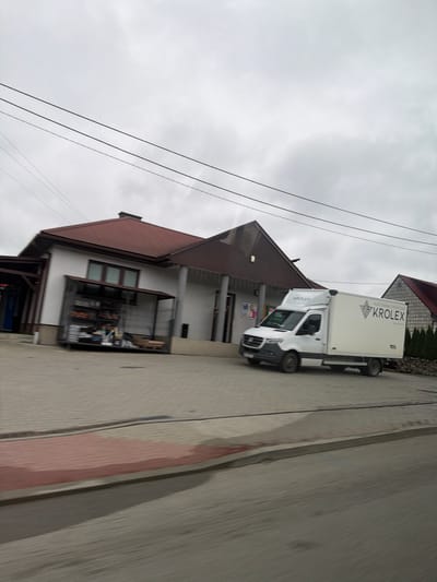 The image shows a white delivery van with "KROLEX" written on its side, parked in front of a building. The van appears to be a Mercedes-Benz Sprinter. To the left of the van is a small building with a red roof, and a metal cage containing shelves with various items. The scene is on a paved area, with power lines overhead. The location is identified as Wilkowisko, Poland. The image is slightly blurred due to motion, suggesting it was taken from a moving vehicle. The sky is overcast.