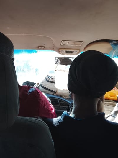 Inside a vehicle, a person wearing a black hat sits in the passenger seat. Bags and luggage are piled up on the dashboard, obscuring the view through the windshield. The car's interior is visible, including the ceiling, rearview mirror, and dashboard vents. The time displayed on the car's digital clock is 20:36. A person wearing black sits in the back, and only the back of his head is visible.