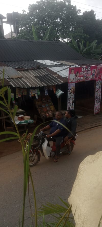 This image captures a street scene in Ezinifite, Nigeria. In the mid-ground, a red and white motorcycle, loaded with a cluster of bananas in a front basket, travels from left to right on a paved asphalt road. The driver is a male wearing a dark blue polo shirt and dark pants, operating the vehicle. A female passenger, dressed in a dark jacket and dark pants, sits behind him.
In the background, a single-story commercial building stands with a weathered corrugated metal roof, showing varied states of repair. The left section of the building functions as an open-fronted stall, displaying numerous packaged goods, sachets, and small items hanging and arranged on wooden racks. A white sack and bowls with produce are visible on a table in front of this stall. The right section of the building features a prominent pink signboard for "O-Z BEAUTY SALON," advertising services with images of women. The salon entrance is a dark opening beneath this sign.
The upper background is dominated by dense green foliage from large trees, with an overcast sky visible above. A black water tank on a metal stand is partially visible above the trees on the far left. Green foliage from an unseen plant or bush partially obscures the immediate foreground of the image.