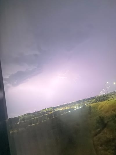 The image captures a dramatic lightning strike illuminating the sky over a cityscape, likely at dusk or night. The lightning forks across the purplish-grey sky, creating a bright, almost ethereal glow. Below, the city lights are visible, suggesting a built-up area. A dark foreground element, possibly a window frame or barrier, partially obstructs the view on the left side of the image. The rest of the scene shows a landscape with some greenery and streetlights lining what appears to be a road or an elevated infrastructure, with a fence or barrier running along the side. The overall impression is one of a powerful natural phenomenon occurring over a populated area.