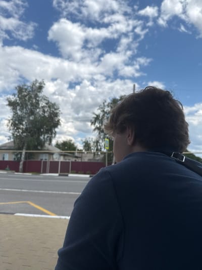 The image is a close-up, side-profile shot of a young person, seen from the shoulder up, with their head tilted downwards. They are wearing a dark blue hooded sweatshirt, with a silver buckle visible near the shoulder. The person's hair is dark brown and appears somewhat tousled. The background is a bright blue sky filled with fluffy white clouds. To the right of the person, and slightly out of focus, is a street scene. Visible elements include a road with white and yellow markings, a light-colored sidewalk or pavement, a dark red fence, and some buildings with trees in front of them. A yellow street sign is also discernible in the mid-ground. The overall impression is of an outdoor setting on a clear, sunny day. No specific activity or event is clearly depicted, but the person's posture suggests they might be looking at something on the ground or engaged in thought. The time of day appears to be daytime, given the brightness of the sky. The location is identified as Shulhynka, Ukraine.