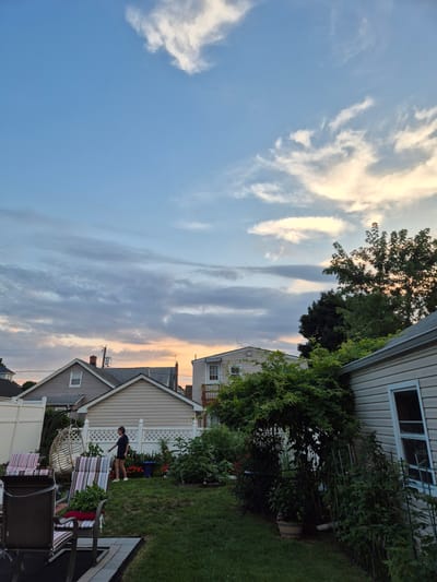 In South River, USA, this image captures a backyard scene during what appears to be sunset. The sky is a gradient of blue, with scattered white and grey clouds, transitioning to a soft orange hue near the horizon. Several houses with pitched roofs are visible in the background, some with windows and chimneys. A person, appearing to be a young woman wearing a dark t-shirt and shorts, is walking across the grassy lawn. In the foreground, outdoor furniture, including chairs with striped cushions and a hanging pod chair, are arranged on the grass and a paved patio area. Lush green vegetation, including bushes, vines climbing on a trellis, and plants in pots, occupies a significant portion of the yard. A white fence separates the backyard from what appears to be a neighbor's property. The overall atmosphere is peaceful and domestic.