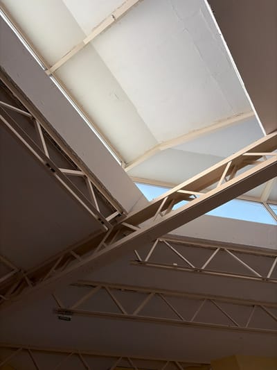 An upward view reveals a building's structural elements. White, triangular ceiling panels are intersected by a skylight showing blue sky. The construction features steel beams with diagonal supports, arranged in a complex grid. The light contrasts with the shaded areas on the ceiling.