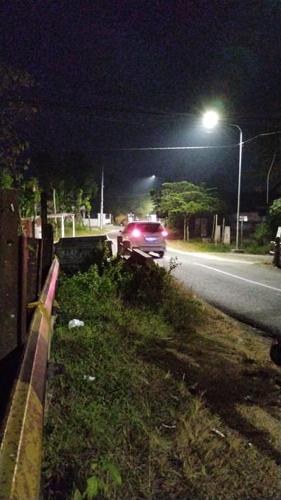 At night, a light-colored car is driving on a paved road in Tulungrejo, Indonesia. The car's brake lights are illuminated. Streetlights illuminate the road and surrounding vegetation. The area appears to be a rural or suburban setting, with trees and some structures visible in the background. The foreground features a grassy embankment next to a yellow and pink barrier. There is a white plastic bag on the grass.