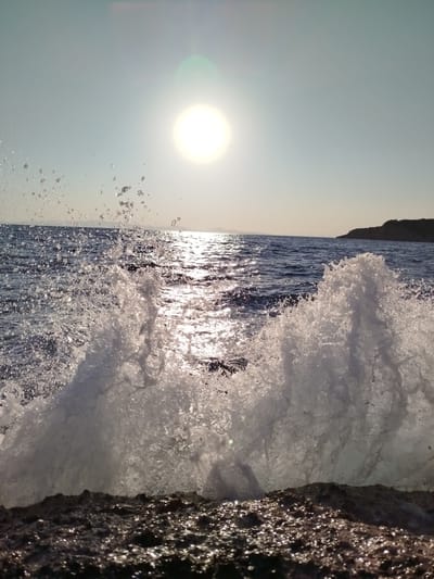 A wave crashes against a rocky shore in Karaburun, Turkey. The sun is low in the sky, casting a golden glow on the water and creating a lens flare in the upper center of the image. The wave is captured in mid-air, with water droplets spraying in all directions. The rocky shore is dark and textured, with some areas appearing wet and shiny. The sea is a deep blue, with gentle waves rolling in the distance. A distant shoreline with a dark, hilly landscape is visible on the right. The image is taken from a low angle, emphasizing the power and drama of the crashing wave.