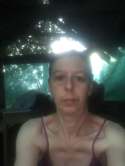 This is a sideways, close-up, selfie-style photograph of a fair-skinned woman with light brown hair and brown eyes. She is wearing a purple strap top. Her expression appears neutral. She is looking at the camera. The background is blurred, suggesting it is out of focus. The lighting is bright and appears to be natural daylight, possibly from a window or outdoors, creating a somewhat blown-out effect in the upper part of the image. The overall scene is dark with a significant bright light source behind the subject. The image quality is grainy and lacks sharp detail.