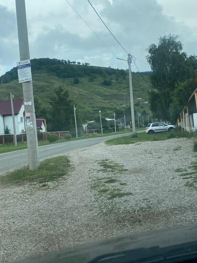 The image depicts a roadside scene in the city of Shiryaevo, Russia, under an overcast sky. A prominent concrete utility pole stands to the left, adorned with a "Rostelecom" advertisement and a phone number, "8 996 79196 29". Adjacent to the pole is a white residential building with a maroon roof, bordered by a wooden fence. The road is paved with asphalt and appears to be a two-lane street. To the right of the road, a white hatchback car is parked on a grassy area. Further in the background, a gently sloping green hill is visible, dotted with trees and some scattered houses. The foreground consists of a gravel and grass area. The overall impression is of a quiet, rural or suburban setting.