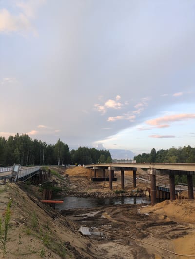 A wide-angle, landscape photograph taken from a low angle shows a bridge under construction spanning a narrow body of water. The bridge structure, made of concrete and supported by large cylindrical pylons, extends from left to right across the frame. The surrounding area is a construction site, with mounds of sand and dirt, muddy patches, and various construction materials like pipes and metal supports visible.
In the foreground, a dirt embankment slopes down towards the water, with sparse green vegetation growing on it. A temporary pedestrian bridge, made of metal, is visible on the left side of the frame, leading towards the main bridge structure. The water appears somewhat murky and reflects the sky above.
The background is dominated by a dense line of green trees, forming a forest that stretches across the horizon. The sky is a mix of light blue with scattered, wispy clouds, some tinged with pink from the setting or rising sun. The overall impression is one of ongoing development in a semi-natural environment.
The location is посёлок Молочной фермы, Russia. The scene depicts a construction site for a new bridge, indicating infrastructure development in the area. There are no people visible in the image. The time of day appears to be either early morning or late evening, given the soft lighting and the hues in the sky.