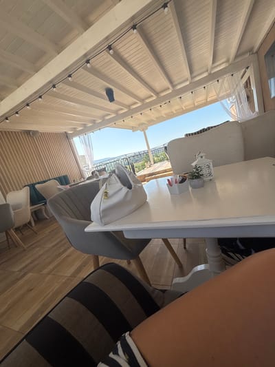 This is a medium shot, taken from a low angle, of a table at an outdoor cafe in Ognyanovo, Bulgaria. The table is white and has a white lantern with decorative cutouts, a small potted plant, and a white container with packets of sugar and artificial sweeteners. A white handbag with a yellow charm hangs from a chair. In the background, there are several chairs and a view of the sky and distant landscape. The ceiling is made of white wooden beams and panels, with string lights. The floor is tiled with a wood-like pattern. The person whose legs are visible in the foreground is sitting on a striped cushion.