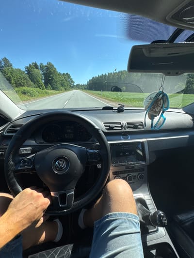 The image shows the interior of a car being driven on a sunny day. The perspective is from the driver's seat, looking forward and slightly to the right. The driver's hands are visible on the steering wheel, and their legs are in denim shorts, indicating warm weather. The dashboard displays a speed of 100, the time as 15:44, and the temperature as 24.5 degrees Celsius. A car's infotainment system is on, playing music by "Gym Class Heroes." The view through the windshield reveals a straight road bordered by green trees and a clear blue sky, suggesting a rural or highway setting. A pair of blue sunglasses hangs from the rearview mirror, along with a circular air freshener. The gear stick, a manual six-speed, is visible to the right of the driver. The scene depicts a leisurely drive in a European setting, likely Latvia given the location context.