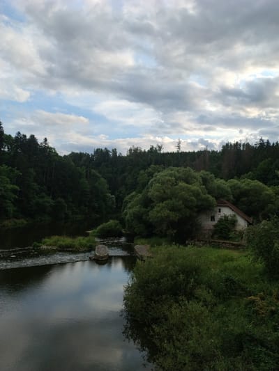 This image captures a serene landscape in Stádlec, Czechia, featuring a river flowing through a lush, forested valley. In the foreground, the calm water reflects the cloudy sky and surrounding greenery. A small dam or weir creates a gentle cascade across the river. On the right bank, nestled among dense trees, stands an old, weathered house with a red-tiled roof and visible windows. The backdrop is a dense wall of trees, characteristic of the Czech countryside, with a dramatic, cloud-filled sky overhead. The scene evokes a sense of peaceful isolation and natural beauty.
