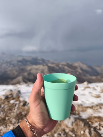 A person's right hand, wearing a silver chain bracelet and a blue jacket sleeve, holds a light green cup containing a greenish beverage. The scene is set outdoors in a mountainous, snow-dusted, rocky landscape, identified as Mali Mikulići, Montenegro. Distant peaks are visible under a cloudy, overcast sky, suggesting cool or inclement weather conditions.