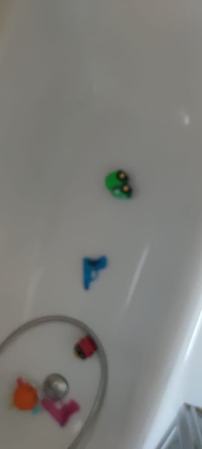 The image shows a close-up, out-of-focus view of what appears to be a bathtub or sink with several small toy items. In the upper right quadrant, there are two green, frog-shaped bath toys. Below that, in the center, is a blue toy gun. In the lower left quadrant, there is a circular object that appears to be a shower head or faucet with a flexible hose. Attached to the hose are several small toys: a red and black toy, an orange toy, a silver spherical object, and a pink object shaped like a "V". The background is a smooth, white surface, typical of a bathroom fixture. There is no indication of the location in Liebenburg, Germany.