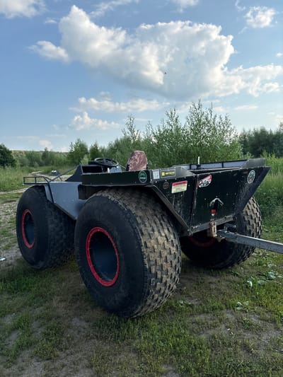 The image shows an all-terrain vehicle (ATV) with oversized tires, possibly a custom-built amphibious vehicle, parked on a grassy, dirt track. The ATV is black with red accents on the wheels. It appears to have a steering wheel and a simple open-air cockpit. There are several stickers on the back of the ATV, some with Cyrillic text. One sticker reads "ВЫСОКО," and another appears to say "ВНИМАНИЕ." The vehicle is positioned at an angle, with the sky and trees in the background. The sky is blue with scattered white clouds, suggesting it is daytime with fair weather. The surrounding environment includes green trees and vegetation, indicative of an outdoor, possibly rural or wilderness setting. The ATV is the primary subject, and there are no people visible. The overall impression is one of a robust, off-road vehicle prepared for challenging terrain. The presence of Cyrillic text on the stickers suggests a location within a Russian-speaking region, consistent with the provided location context of Drakino, Russia.