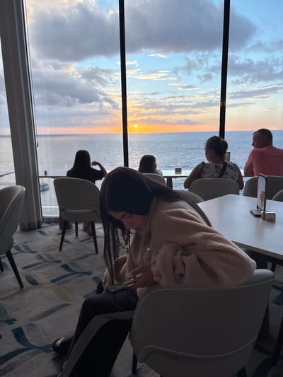 The image depicts an interior scene with a panoramic ocean view at sunset, observed from what appears to be a lounge or restaurant situated in the city of San Miguel de Cozumel, Mexico. The location offers a westward vista over the Caribbean Sea.
In the foreground, a young female with long, dark hair is seated in a light-colored chair, engrossed in a white smartphone held in her hands. She wears a light beige, textured fleece-style jacket and dark pants with a lighter stripe down the outer leg. Her gaze is directed downwards at the device.
Beyond her, and framed by large, dark-paned windows, four other individuals are visible, primarily from behind, seated and facing the ocean. From left to right: a person with dark hair and a dark top, a partially obscured person with dark hair, an adult female wearing a patterned light-colored top, and an adult male in a light pink collared shirt, seen in right profile. All individuals facing the windows appear to be observing the external environment.
The interior features modern, light-colored chairs and white rectangular tables, one of which holds a menu display. The floor is carpeted with an abstract pattern in shades of blue, green, and gray.
Outside the windows, the sun is depicted as a bright, orange-yellow orb, low on the horizon, setting over the calm, dark blue ocean. The sky above is a mix of scattered clouds, illuminated with orange and pink hues near the horizon, transitioning to blue and gray higher up. A linear structure, resembling a pier or jetty, extends into the water on the far left of the distant view.