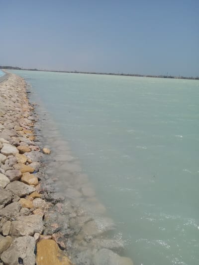 The image depicts a stretch of water along a rocky shoreline, under a clear sky. The water is pale blue-green and appears slightly choppy, indicating a breeze. The shoreline is constructed of large, irregularly shaped rocks, ranging in color from gray to light brown. In the distance, a coastline or breakwater is visible, with a faint skyline suggestive of buildings or structures. The location is Al Khobar, Saudi Arabia, based on the context of the prompt. There are no people visible in the frame. The scene appears to be a man-made or modified coastal environment.