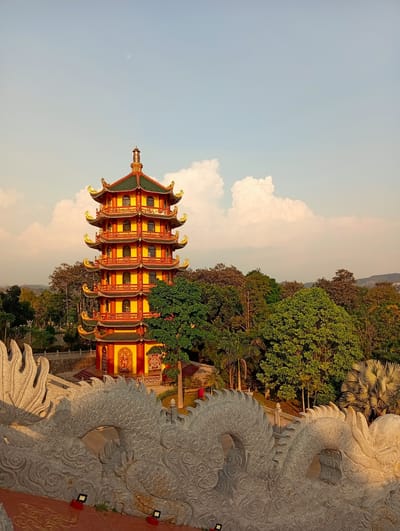 This elevated, wide shot captures a vibrant, multi-tiered East Asian-style pagoda set amidst a lush, forested landscape under a serene sky, located in Phú An, Vietnam. The pagoda, the central subject, is strikingly colored in red and yellow with distinctive green roofs and elaborate golden accents on its upturned eaves. It features multiple levels, each with balconies and windows, showcasing intricate architectural details.
The pagoda is surrounded by a dense canopy of various green trees, creating a verdant environment that stretches towards a hazy horizon. The sky is a gentle blue with scattered white cumulus clouds, and a subtle, faint moon is visible in the upper portion, suggesting the warm, golden light of either late afternoon or early morning.
In the immediate foreground, a large and intricately carved stone balustrade shaped like a dragon spans the lower part of the frame. Its detailed scales and wave-like textures are illuminated by the warm, low-angle sunlight. Beyond this stone dragon railing, and partially visible to the left of the main pagoda, is a smaller, red and yellow structure with a golden statue prominently displayed in an alcove, indicating a larger temple complex.
No people are visible, and no specific event or activity is taking place, allowing the focus to remain on the majestic architecture and natural surroundings. There is no discernible text visible in the image.