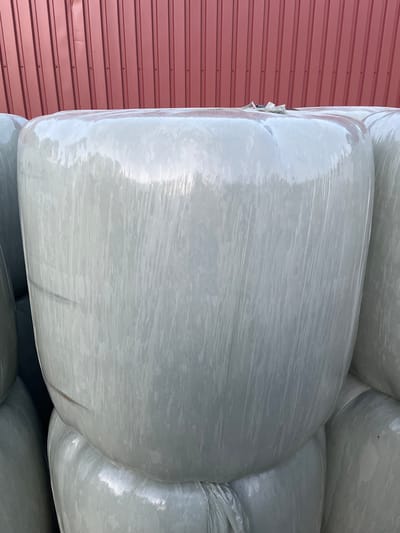 This image shows a close-up of several large, round bales wrapped in white plastic, stacked in front of a red corrugated metal wall. The bales appear to be hay or silage, commonly used for animal feed. The plastic wrapping is somewhat translucent and shows creasing and wrinkles from the wrapping process. In the upper center of the main bale, a small, crumpled piece of paper or plastic is visible, possibly a tag or marker. The red wall behind the bales has vertical lines and appears to be a building facade. The scene is outdoors, with natural lighting creating highlights and shadows on the plastic. The context of Uithuizen, Netherlands, suggests this is likely a rural setting, perhaps a farm or agricultural storage area.